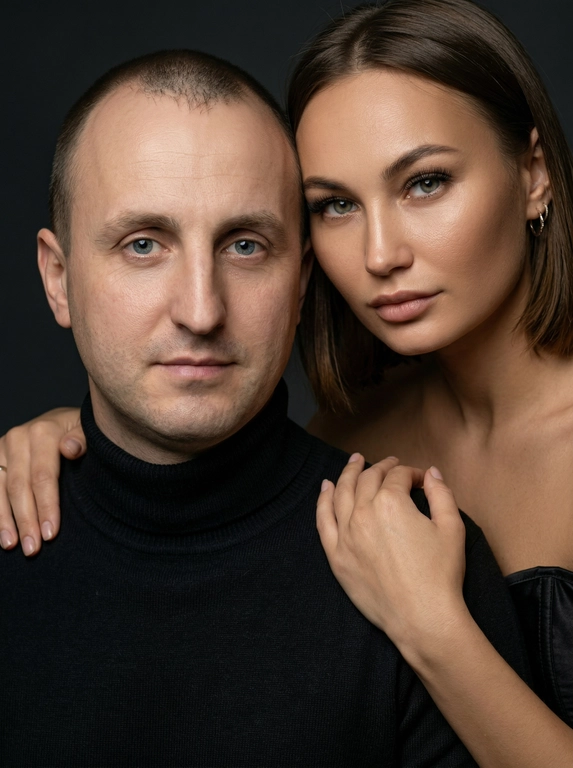 Гиперреалистичный студийный fashion-портрет молодой пары по загруженному референсу, 100% сходство лиц. Черты лица не изменять. Крупный план, кадр по плечи. Мужчина, спокойный уверенный взгляд прямо в камеру, естественное выражение лица, одет в черный трикотажный свитер с высоким воротом. Женщина стоит позади мужчины и нежно обнимает его, ее руки лежат на его плечах. Ее лицо расположено рядом с его лицом, она повернута слегка боком, голова немного наклонена вперед, взгляд направлен прямо в камеру. У женщины выразительные глаза с длинными густыми черными ресницами, естественный clean-glam макияж, натуральные брови, теплый оливковый оттенок кожи, небольшие серьги-кольца. Композиция плотная и интимная, их лица расположены очень близко друг к другу, современный модный портрет пары. Женщина одета в черный корсет. Кожа максимально натуральная: живая текстура кожи, видимые микропоры, subtle skin glow, естественный мягкий блеск кожи, natural skin microtexture, skin translucency, micro skin details, легкие естественные несовершенства кожи, без ретуши и без эффекта пластика. Освещение: мягкий студийный beauty lighting, рассеянный ключевой свет спереди и немного сбоку, мягкие глубокие тени для объема лица, деликатные световые блики на коже. Фон: глубокий темный студийный фон, темно-графитовый или почти черный, создающий контраст с кожей и лицами. Стиль: luxury fashion editorial photography, high fashion portrait, ultra photorealistic, 85mm portrait lens, shallow depth of field, creamy bokeh, cinematic soft lighting, RAW photo, extremely detailed, fine hair strands, natural skin pores, professional photography, photorealism, 8k, high dynamic range.