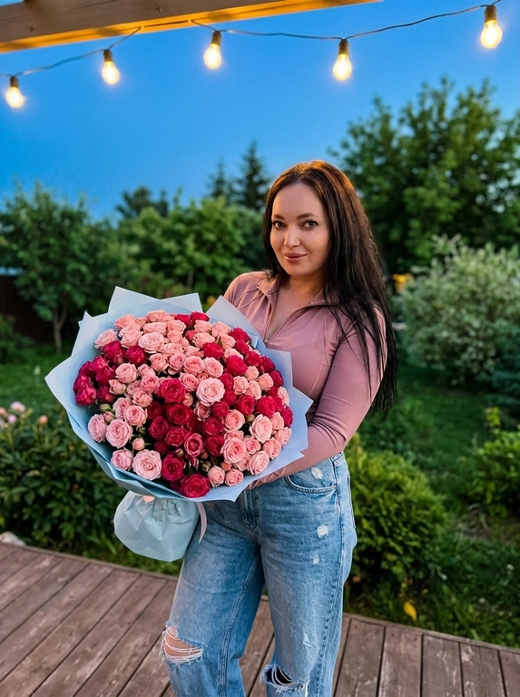 Фотореалистичный снимок модели в полный рост, стоящей на террасе. Она держит в руках огромный, невероятно пышный букет. Букет завернут в светло-голубую упаковочную бумагу. Модель смотрит прямо в камеру с мягкой, немного застенчивой мягкой улыбкой. 
На ней нежно розовый спортивный лонгслив на замке слегка расстёгнутый,
с длинными рукавами,светло голубые свободные джинсы с потертостями. Задний план представляет собой зеленый сад с деревьями и кустарниками. Над ней видна гирлянда с теплыми лампочками, которые создают уютное освещение и красивые блики. Небо имеет глубокий синий оттенок. Освещение смешанное: мягкий естественный свет вечера и теплый искусственный свет от гирлянды, что создает романтичную, сказочную и очень теплую атмосферу.
Фото в золотой час и кинематографический эффект с добавлением зерна.
Стиль RAW.

Какие будут цветы?: В руках из розовых кустовых роз разных оттенков — от нежно-розового до насыщенного малинового.