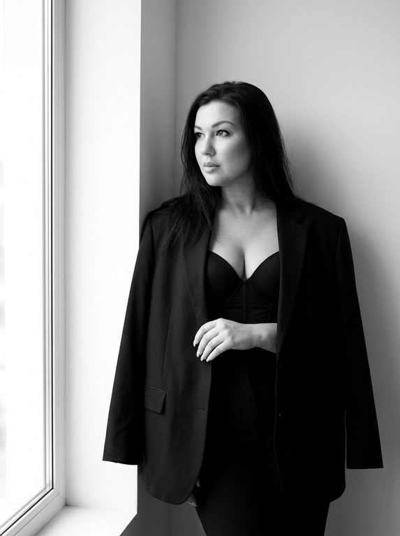 Женщина слегка наклонилась к стене в углу комнаты. Сохрани лицо и черты лица с оригинального фото. На ней надет большой чёрный пиджак, накинутый на плечи, и чёрный лаконичный топ-корсет с V-образным вырезом. Женщина стоит, смотря в сторону окна вдаль, выражение лица спокойное и уверенно задумчивое. Она слегка опирается на стену, положив руку на пиджак. Комната светлая, с большими окнами слева, пропускающими естественный свет. Общая атмосфера спокойная и стильная, подчёркивающая элегантность и минимализм. Положение женщины в кадре — справа от центра. Освещение мягкое, естественное, создающее ощущение минимализма и простоты. Фотография выполнена в чёрно-белом стиле, подчёркивающем контрасты и текстуры. Атмосфера спокойная, элегантная и немного загадочная. Максимально близкий ультрареалистичный 8K портрет.