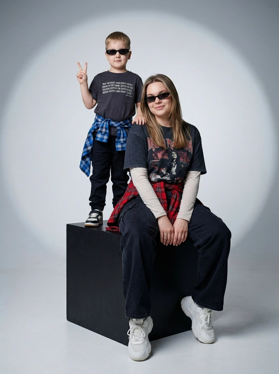 Photorealistic studio portrait photography in minimalistic fashion/editorial aesthetics, static camera at chest level of a seated person, light frontal angle without noticeable perspective deformation; composition with two characters: an model sits on a large matte black cube, body slightly turned towards the camera, knees bent, feet on the floor, the arms are lowered and relaxed in front of the knees, the fingers are neatly extended; the child stands on the same cube behind and to the left, at full height, one hand raised with a "V" gesture, the other lowered, pose confident and playful; both wearing narrow dark sunglasses, gaze directed at the camera; shooting plan — full height for both with a small supply of air above and below, center the center of the frame is shifted to the right to a sitting figure, a standing child balances on the left; the lens is normal/short portrait 50-70 mm, without width; aperture is approximately f/5.6–f/8, moderate depth of field, both characters are sharp, the background is slightly soft; the studio lighting is soft, with one key source on the front left at a level just above the head (softbox), giving smooth transitions and a pronounced but soft shadow against the background on the right; light filling light from the front reduces contrast, shadows are readable; the background is a smooth, light, seamless cyclorama/wall with a noticeable large oval light spot (gradient) in the center, there is a darker vignetting around, a clean minimalistic studio without props; the mood is calm, stylish, slightly retro, with an emphasis on street fashion and family vibe; the expression on the face of the seated model is a soft smile, relaxation; the person standing has a confident neutral facial expression; clothes: the seated model has a dark oversized T-shirt with a large rectangular print on the chest, under it a light longsleeve with long sleeves, wide dark cargo trousers/joggers with pleats at the knees and bottom, a plaid shirt/flannel tied at the waist with a knot, massive light sneakers with a pronounced sole and mesh texture; the child has a dark T-shirt with a large inscription on the chest, dark trousers, a plaid shirt tied at the waist, sneakers in two colors; fabrics with a realistic cotton and flannel texture, noticeable fine folds, natural creases on the knees and in the waist area, matte surfaces without gloss; micro-details: natural skin texture without plastic, neat glare on the lenses of glasses, a clear contour of the shadow from the raised hand on the background, clean floor and soft drop shadow from the cube; high detail, natural sharpness, light grain like a film shot, moderate contrast, soft matte/film look, no HDR, no overexposure and no unnecessary objects.

Какого цвета будут рубашки на поясе?: Красный и синий.