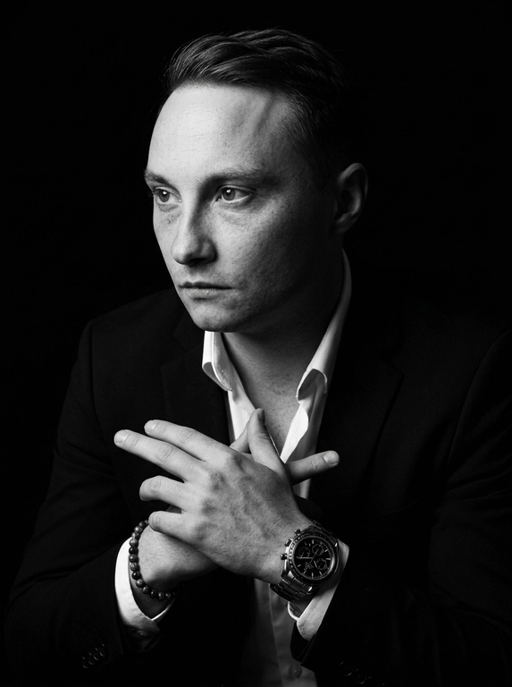 A hyper-realistic 8K cinematic portrait of a uploaded person,
captured in a dramatic Chiaroscuro style. The subject is seated at a
three-quarter angle, leaning slightly forward with a relaxed yet
commanding posture. His face is turned away from the lens, with
one side of his face clearly illuminated while the other is
completely lost in deep, velvety black shadows. His expression is
contemplative, not looking at the camera. The pose is precise: his
hands are clasped together near his chest; one hand is held
straight with fingers slightly tilted, while the other hand is
positioned so only the fingers are visible, resting over the first. He
wears a luxury black chronograph watch with a detailed metal link
bracelet, and a beaded bracelet. His attire is a sharp
black suit jacket over a white dress shirt with the top buttons
open, showing refined fabric textures. The background is a solid,
seamless black. The lighting is sharp and directional, highlighting
the intricate details of his hair,
the moisture in his eyes, and the glint off the watch face.
Cinematic black-and-white color grade with rich contrast and
smooth, natural skin transitions. Shot on 85mm lens, f/1.8, high-
resolution commercial photography