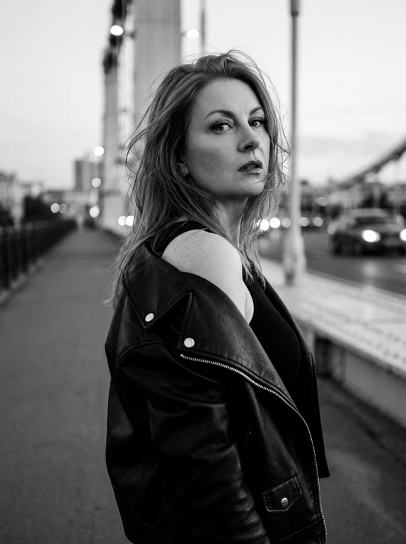Используй внешность модели с фото. Фотореалистичное черно-белое фото в стиле модной редакционной съемки с эстетикой пленочной фотографии (зернистость, высокий контраст). Модель с волосами слегка растрепанными ветром. Она одета в черную, объемную кожаную куртку-косуху, которая небрежно спущена с левого плеча, открывая его. Под курткой надет черный облегающий топ. Поза: модель стоит вполоборота спиной к камере, но резко оборачивается и смотрит назад через плечо прямо в объектив. Выражение лица серьезное, дерзкое и чувственное, губы слегка приоткрыты, густые ресницы. Фон размыт, видны элементы конструкции моста и огни проезжающих машин вдали, создающие городскую атмосферу. Освещение естественное, подчеркивающее текстуру кожи и блеск кожи куртки. Высокое разрешение, 8k. Освещение мягкое и равномерное, без жестких теней, свет подчеркивает блеск жемчуга и страз, цвета насыщенные, но реалистичные. Высокая детализация, фотореализм, четкий фокус на лице, естественные пропорции.
