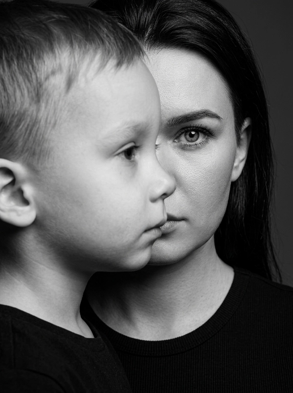 Чёрно-белый фотореалистичный студийный beauty-fashion портрет родителя и ребёнка с представленных фото. Крупный плотный кадр по плечи, без свободного пространства. Ребёнок находится на переднем плане, ближе к камере, в профиль, расположен сбоку и частично перекрывает лицо родителя. Лицо ребёнка закрывает примерно половину лица родителя, создавая сильный эффект визуального перекрытия. Родитель находится позади ребёнка, строго по центру кадра; у родителя виден только один глаз, направленный прямо в камеру, вторая часть лица скрыта лицом ребёнка. Лица расположены максимально близко, почти соприкасаются, но без явного физического действия. Ребёнок смотрит в сторону, выражение спокойное, сдержанное и закрытое. Родитель смотрит прямо в камеру одним открытым глазом, взгляд холодный, спокойный и пронизывающий. Атмосфера high-fashion editorial: минимальная эмоциональность, люксовая отстранённость, скрытое напряжение и холодная близость. Фон тёмный, чистый, студийный, без деталей. Жёсткое студийное освещение, высокий контраст, глубокие тени, выразительная светотень на лицах, натуральная текстура кожи, sharp focus, dramatic black and white photography, editorial beauty portrait, без улыбок, без лишних предметов, без текста и логотипов.