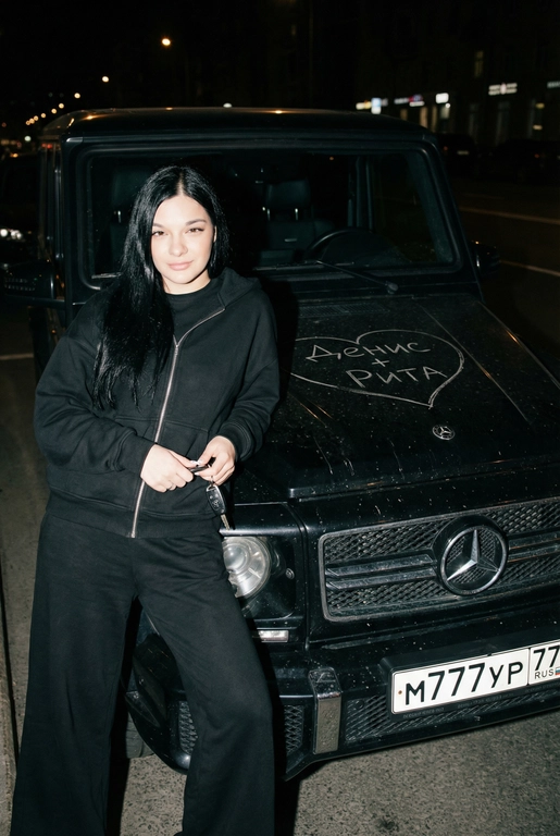 Строго сохранить лицо с прикреплённой референс-фотографии, на 100% без искажений. Человек одет полностью в чёрный, монохромный оверсайз спортивный костюм: свободная худи на молнии и широкие расслабленные штаны, без логотипов, текста и брендинга. Акцент на реальной текстуре кожи и детализированной текстуре ткани.
Сырой кинематографичный ночной кадр. Человек опирается на капот автомобиля, средний план, лёгкий верхний ракурс. В одной руке игриво держит ключи от машины. Номерной знак виден: m777yp 77.
Строго по центру капота, покрывая всю поверхность капота, глубоко и грубо процарапаны (не мелом) царапины: большое сердце с текстом «» внутри.
Жёсткая прямая вспышка, сильные тени, высокий контраст, видимая пыль и микро-царапины на краске. Тёмный размытый городской фон.
Объектив 35mm, f/2.8, грубая уличная эстетика.

Надпись на капоте: Денис + Рита 

Марка автомобиля: Опирается на чёрный Mercedes-Benz G-Class