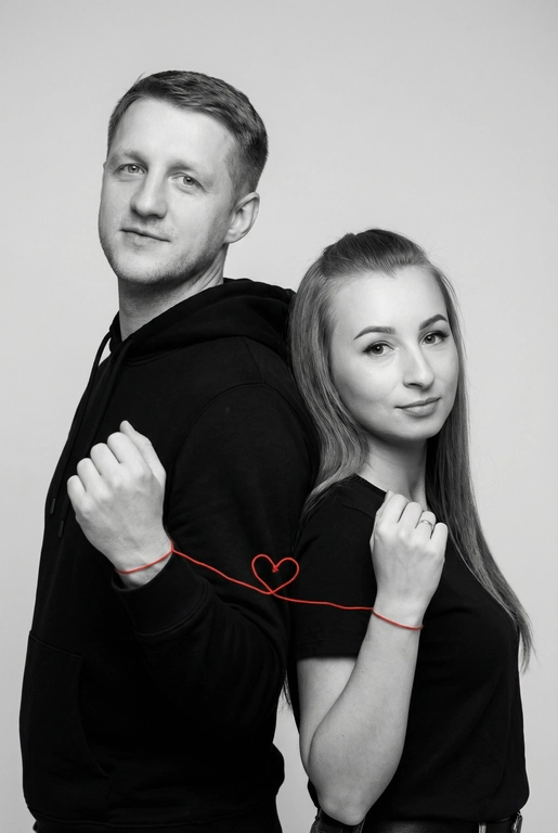 Фотореалистичный чёрно-белый студийный портрет пары, стоящей спина к спине, слегка повернувшись к камере. Оба в минималистичных чёрных образах (худи и футболка), с мягкими уверенными выражениями лица, естественный внешний вид. Тонкая красная нить, связанная вокруг их запястий, соединяет их и образует маленькое сердце между ними. Чистый светло-серый студийный фон, мягкое рассеянное освещение, высокая детализация текстуры кожи, резкий фокус, кинематографичная композиция, вертикальный формат 9:16, минималистичная эстетика, высокое разрешение, профессиональная фотография.

