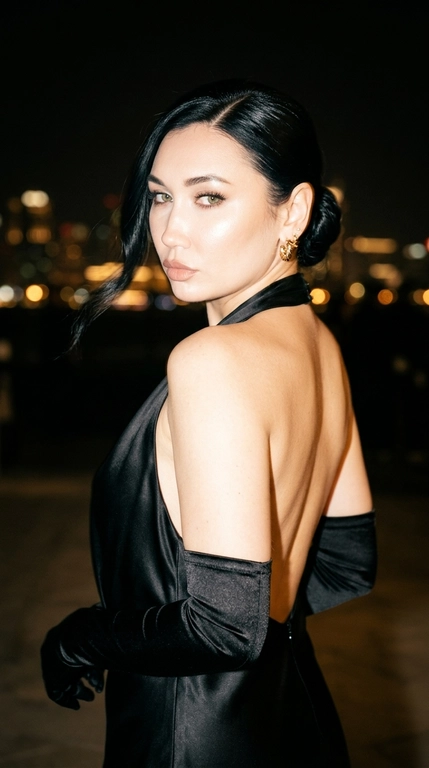 Фотореалистичный luxury glamour editorial. Крупный портрет по пояс. Женщина стоит почти спиной к камере и в момент съёмки резко оборачивается через плечо, словно случайно заметила фотографа. Голова находится в лёгком движении, создавая естественный динамичный кадр без постановочности. Взгляд направлен прямо в объектив — спокойный, холодный и уверенный.

Главный акцент — роскошная вечерняя эстетика, эффект случайно пойманного кадра и выразительная динамика поворота. Один глаз может быть частично или полностью прикрыт волосами, второй остаётся главным акцентом кадра — выразительный, глубокий взгляд с сильным зрительным контактом с камерой.

Макияж — luxury red carpet glam: безупречная кожа с естественным сатиновым свечением, деликатный контуринг, мягкий золотистый хайлайтер на скулах и во внутренних уголках глаз, выразительный смоки с вытянутой графичной стрелкой к виску, затемнение межресничного пространства для глубины взгляда, очень густые длинные ресницы с драматичным объёмом, естественные объёмные брови с чёткой архитектурой, естественные губы цвета капучино с мягким сатиновым объёмом и идеально прорисованным контуром.

Фон — глубокая ночная темнота с едва заметными, сильно размытыми тёплыми огнями города, без людей, без красной дорожки, без фотографов и без лишних объектов. Атмосфера дорогого вечернего мероприятия.

Освещение — мощная встроенная вспышка прямо в объектив. Качество снимка намеренно имитирует дорогую компактную цифровую мыльницу конца 90-х — начала 2000-х: жёсткая фронтальная вспышка, слегка пересвеченные блики на коже, характерный CCD-цифровой шум, лёгкая хроматическая аберрация, мягкая цифровая резкость, небольшое виньетирование, натуральные тёплые цвета, отсутствие HDR, лёгкий motion blur от движения модели и камеры, ощущение случайно пойманного кадра.

Цветокоррекция минимальная — естественная цифровая фотография начала 2000-х, без современной журнальной обработки и без пластиковой ретуши и без людей на фоне.

Какая будет прическа?: Волосы гладко собраны в низкий плотный пучок у основания шеи с глубоким боковым пробором. Одна длинная мягкая прядь выпущена у лица и слегка движется от поворота головы. Остальная масса волос идеально собрана, подчёркивая открытую спину, серьги и линию шеи. Укладка выглядит дорого, строго и по-красно дорожному. Волосы покрыты тончайшей невесомой вуалью лака с мельчайшими золотистыми микроблёстками

Какая будет одежда?: На женщине роскошное чёрное вечернее платье из плотного струящегося сатина с полностью открытой спиной и высоким воротом-халтер. Платье плотно облегает верхнюю часть корпуса и выглядит элегантно, строго и очень дорого. Ткань красиво ловит вспышку, создавая мягкие блики и подчёркивая силуэт. Образ дополняют длинные чёрные атласные перчатки выше локтя.