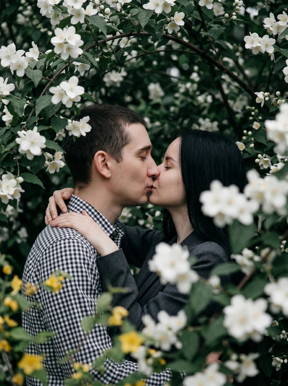 A man and a woman stand face to face, kissing—their lips touching, faces in profile, eyes closed. The shot is close-up, from shoulders to head, angled at eye level, with their faces centered in the frame and surrounded by flowering branches. The foreground and edges of the frame are filled with out-of-focus branches and flowers. Do not alter the faces from the uploaded photos.
The skin appears perfectly smooth and matte.
The man is wearing a cotton shirt with a small gingham check in black and white tones. The woman is wearing a dark gray jacket.
The location is a flowering tree with dense branches: large white flowers with five petals and yellow stamens —presumably jasmine or bird cherry—and dense dark green foliage. The branches frame the figures on all sides, creating a living, arched tunnel. In the left middle ground, yellow flowers are out of focus.
The lighting is natural, overcast, diffused, with no clear direction of the light source—an even, soft light. Shadows are virtually absent. Contrast is low. The overall tone of the frame is muted and cool, with green and white dominant tones. The highlights on the skin are subdued and matte. The film grain is pronounced.
Shot with an 85mm lens, minimal depth of field with sharp focus exclusively on the lips and the area where the faces meet—the foreground with the man's shoulder and the nearby branches are completely out of focus, the background with foliage is blurred into bokeh. Pronounced film grain. A reportage paired shot in the style of a Candid Film portrait, highly detailed and photorealistic.
Soft romantic spring botanical mood,
dominant tones: white blossom, deep green, dark charcoal,
low saturation with cool green emphasis,
flat diffused overcast natural lighting,
minimal shadow definition,
heavy analog film grain throughout,
lifted blacks with a soft hazy quality,
shallow depth of field with strong foreground blur,
Candid Film Spring color grading.