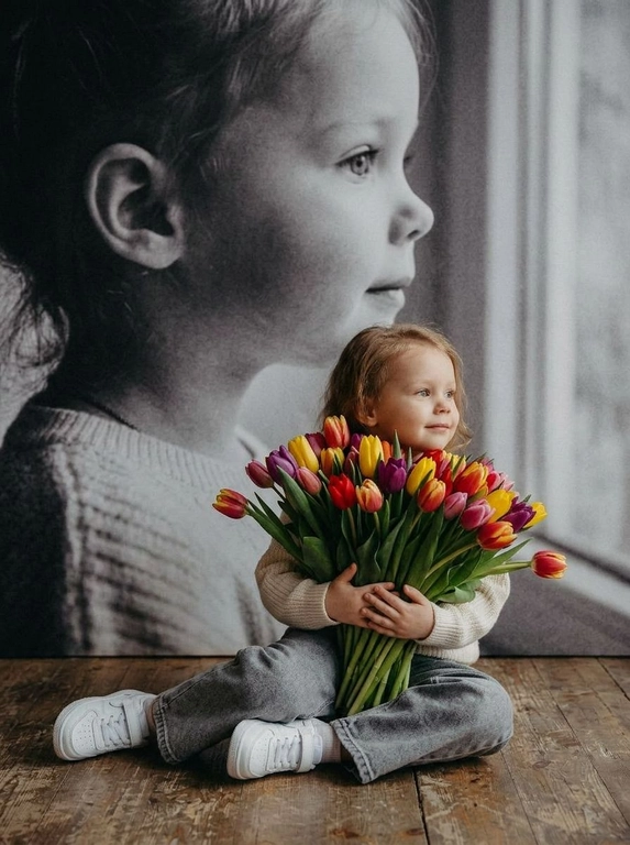 Создай портрет, не меняя черты лица, и превратите его в художественный портрет.
Ребенок сидит на полу в изящной позе, в белой кофте, мягких свободных серых джинсах и белых спортивных кроссовках. Ребенок держит в руках огромный букет ярких разноцветных тюльпанов.
Выражение лица — лёгкая улыбка, ребенок смотрит в сторону. 
Фон — тот же его профиль, в мягком крупном чёрно-белом художественном стиле, крупный план сбоку.
Композиция должна быть художественной, мягкой, реалистичной.
Создай изображение, которое выглядит как настоящая фотография. 
Не меняй черты лица.