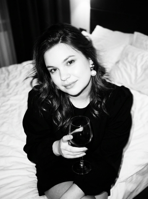 Create a portrait, transfer the face from the reference photo without changing facial features, proportions, or expression, preserve full identity. Aesthetic black-and-white nighttime Y2K photograph captured with direct on-camera flash, shot in motion.
A woman is sitting on the edge of a white, slightly rumpled bed, the frame taken from a slightly elevated angle. She wears a stylish black top with voluminous sleeves and large round statement earrings. In one hand she holds an elegant glass of dark wine. Her body is subtly moving, creating natural motion blur across the face and shoulders, as if the moment was caught mid-gesture. The gaze is languid and directed toward the camera, with a soft, intimate smile. Hair falls in large waves with loose strands drifting across the face, slightly displaced by movement. Lighting is harsh flash against deep night shadows, high contrast with blown highlights and strong falloff, visible film grain. Skin texture remains real and alive, no smoothing, enhanced by flash glare. The mood is high-fashion editorial, Parisian chic, old-money sensuality, nighttime melancholy, spontaneous and cinematic, shot in a raw 35mm film aesthetic with pronounced motion blur and lived-in realism.