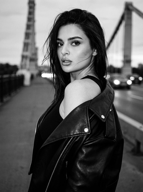 Используй внешность модели с фото. Фотореалистичное черно-белое фото в стиле модной редакционной съемки с эстетикой пленочной фотографии (зернистость, высокий контраст). Модель с волосами слегка растрепанными ветром. Она одета в черную, объемную кожаную куртку-косуху, которая небрежно спущена с левого плеча, открывая его. Под курткой надет черный облегающий топ. Поза: модель стоит вполоборота спиной к камере, но резко оборачивается и смотрит назад через плечо прямо в объектив. Выражение лица серьезное, дерзкое и чувственное, губы слегка приоткрыты, густые ресницы. Фон размыт, видны элементы конструкции моста и огни проезжающих машин вдали, создающие городскую атмосферу. Освещение естественное, подчеркивающее текстуру кожи и блеск кожи куртки. Высокое разрешение, 8k. Освещение мягкое и равномерное, без жестких теней, свет подчеркивает блеск жемчуга и страз, цвета насыщенные, но реалистичные. Высокая детализация, фотореализм, четкий фокус на лице, естественные пропорции.