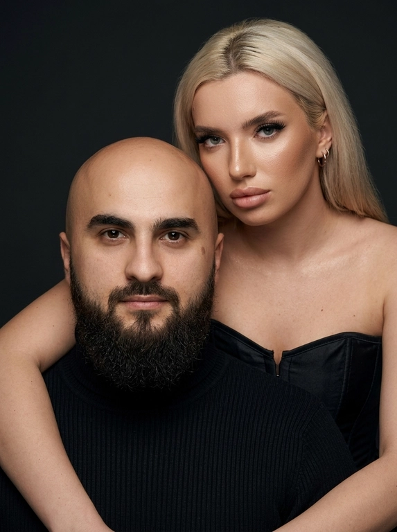 Гиперреалистичный студийный fashion-портрет молодой пары по загруженному референсу, 100% сходство лиц. Черты лица не изменять. Крупный план, кадр по плечи. Мужчина, спокойный уверенный взгляд прямо в камеру, естественное выражение лица, одет в черный трикотажный свитер с высоким воротом. Женщина стоит позади мужчины и нежно обнимает его, ее руки лежат на его плечах. Ее лицо расположено рядом с его лицом, она повернута слегка боком, голова немного наклонена вперед, взгляд направлен прямо в камеру. У женщины выразительные глаза с длинными густыми черными ресницами, естественный clean-glam макияж, натуральные брови, теплый оливковый оттенок кожи, небольшие серьги-кольца. Композиция плотная и интимная, их лица расположены очень близко друг к другу, современный модный портрет пары. Женщина одета в черный корсет. Кожа максимально натуральная: живая текстура кожи, видимые микропоры, subtle skin glow, естественный мягкий блеск кожи, natural skin microtexture, skin translucency, micro skin details, легкие естественные несовершенства кожи, без ретуши и без эффекта пластика. Освещение: мягкий студийный beauty lighting, рассеянный ключевой свет спереди и немного сбоку, мягкие глубокие тени для объема лица, деликатные световые блики на коже. Фон: глубокий темный студийный фон, темно-графитовый или почти черный, создающий контраст с кожей и лицами. Стиль: luxury fashion editorial photography, high fashion portrait, ultra photorealistic, 85mm portrait lens, shallow depth of field, creamy bokeh, cinematic soft lighting, RAW photo, extremely detailed, fine hair strands, natural skin pores, professional photography, photorealism, 8k, high dynamic range.