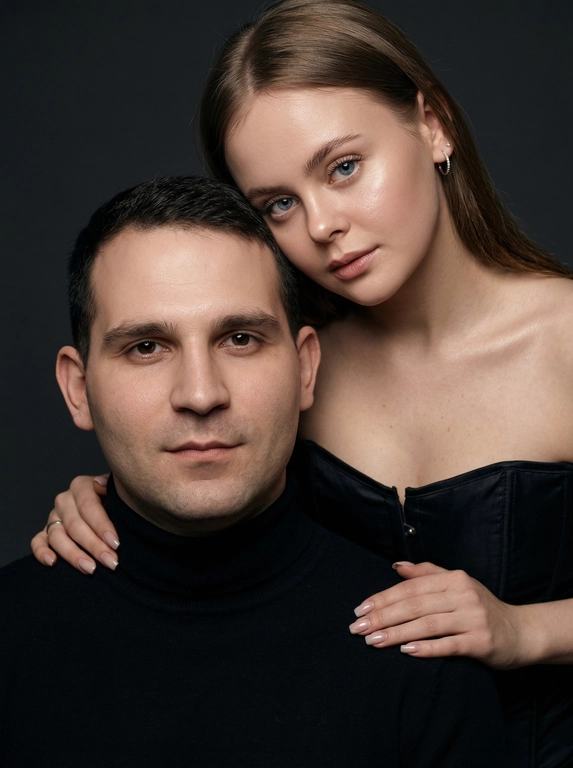 Гиперреалистичный студийный fashion-портрет молодой пары по загруженному референсу, 100% сходство лиц. Черты лица не изменять. Крупный план, кадр по плечи. Мужчина, спокойный уверенный взгляд прямо в камеру, естественное выражение лица, одет в черный трикотажный свитер с высоким воротом. Женщина стоит позади мужчины и нежно обнимает его, ее руки лежат на его плечах. Ее лицо расположено рядом с его лицом, она повернута слегка боком, голова немного наклонена вперед, взгляд направлен прямо в камеру. У женщины выразительные глаза с длинными густыми черными ресницами, естественный clean-glam макияж, натуральные брови, теплый оливковый оттенок кожи, небольшие серьги-кольца. Композиция плотная и интимная, их лица расположены очень близко друг к другу, современный модный портрет пары. Женщина одета в черный корсет. Кожа максимально натуральная: живая текстура кожи, видимые микропоры, subtle skin glow, естественный мягкий блеск кожи, natural skin microtexture, skin translucency, micro skin details, легкие естественные несовершенства кожи, без ретуши и без эффекта пластика. Освещение: мягкий студийный beauty lighting, рассеянный ключевой свет спереди и немного сбоку, мягкие глубокие тени для объема лица, деликатные световые блики на коже. Фон: глубокий темный студийный фон, темно-графитовый или почти черный, создающий контраст с кожей и лицами. Стиль: luxury fashion editorial photography, high fashion portrait, ultra photorealistic, 85mm portrait lens, shallow depth of field, creamy bokeh, cinematic soft lighting, RAW photo, extremely detailed, fine hair strands, natural skin pores, professional photography, photorealism, 8k, high dynamic range.
