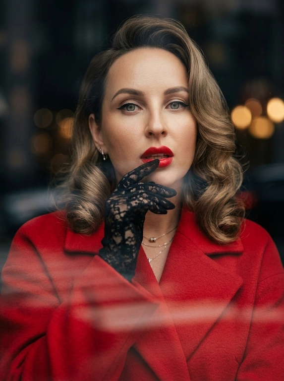 Фотореалистичный fashion-портрет женщины.
Элегантная, уверенная, кинематографичная съемка в стиле high fashion editorial.
Крупный портрет по грудь, легкий поворот корпуса, взгляд прямо в камеру, слегка приоткрытые ярко-красные губы, выразительный макияж с черной стрелкой eyeliner.
На ней ярко-красное пальто с плотной тканью и аккуратным кроем, образ роскошный, дерзкий и женственный.
Одна рука в черной кружевной перчатке поднята к лицу, пальцы слегка касаются подбородка и губ, создавая изысканную позу.
Украшения минимальные, небольшие элегантные серьги.
Съемка через стекло или окно, на переднем плане мягкие размытые отражения и легкие полосы стекла, как у уличной модной фотографии.
Фон темный, размытый, с мягким боке и атмосферой города, но без отвлекающих деталей.
Освещение мягкое, теплое, контрастное, с красивыми тенями на лице, skin glow, естественная текстура кожи, дорогой журнальный стиль.
Цветокоррекция: теплые глубокие оттенки, акцент на красный цвет пальто и губ, благородные темные тени, cinematic mood.
Фото выглядит как дорогая съёмка для модного журнала, ultra realistic, high detail, sharp focus on face, shallow depth of field, 85mm portrait lens, soft film grain, luxury editorial aesthetics.
Негативный промпт
искаженное лицо, другая личность, лишние пальцы, лишние руки, деформированные губы, кривые глаза, плохая анатомия, мультяшность, пластик, чрезмерная ретушь, низкое качество, размытое лицо, шум, пересвет, артефакты, лишние украшения, лишний фон, неестественная кожа, плохой макияж, кривой нос, неправильные пропорции, двойное лицо

Какая будет укладка?: Укладка в стиле ретро-glam: волосы аккуратно уложены в крупные гладкие волны с выраженным объёмом у корней, мягким боковым пробором и глянцевой, ухоженной текстурой. Волны обрамляют лицо и создают элегантный образ в духе классической голливудской съёмки.