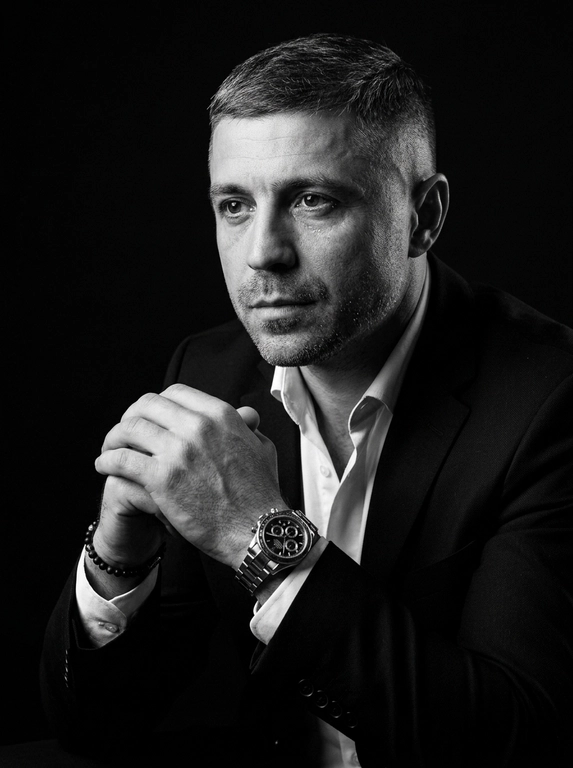 A hyper-realistic 8K cinematic portrait of a uploaded person,
captured in a dramatic Chiaroscuro style. The subject is seated at a
three-quarter angle, leaning slightly forward with a relaxed yet
commanding posture. His face is turned away from the lens, with
one side of his face clearly illuminated while the other is
completely lost in deep, velvety black shadows. His expression is
contemplative, not looking at the camera. The pose is precise: his
hands are clasped together near his chest; one hand is held
straight with fingers slightly tilted, while the other hand is
positioned so only the fingers are visible, resting over the first. He
wears a luxury black chronograph watch with a detailed metal link
bracelet, and a beaded bracelet. His attire is a sharp
black suit jacket over a white dress shirt with the top buttons
open, showing refined fabric textures. The background is a solid,
seamless black. The lighting is sharp and directional, highlighting
the intricate details of his hair,
the moisture in his eyes, and the glint off the watch face.
Cinematic black-and-white color grade with rich contrast and
smooth, natural skin transitions. Shot on 85mm lens, f/1.8, high-
resolution commercial photography