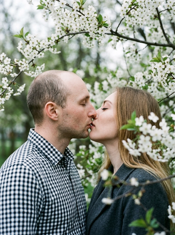 A man and a woman stand face to face, kissing—their lips touching, faces in profile, eyes closed. The shot is close-up, from shoulders to head, angled at eye level, with their faces centered in the frame and surrounded by flowering branches. The foreground and edges of the frame are filled with out-of-focus branches and flowers. Do not alter the faces from the uploaded photos.
The skin appears perfectly smooth and matte.
The man is wearing a cotton shirt with a small gingham check in black and white tones. The woman is wearing a dark gray jacket.
The location is a flowering tree with dense branches: large white flowers with five petals and yellow stamens —presumably jasmine or bird cherry—and dense dark green foliage. The branches frame the figures on all sides, creating a living, arched tunnel. In the left middle ground, yellow flowers are out of focus.
The lighting is natural, overcast, diffused, with no clear direction of the light source—an even, soft light. Shadows are virtually absent. Contrast is low. The overall tone of the frame is muted and cool, with green and white dominant tones. The highlights on the skin are subdued and matte. The film grain is pronounced.
Shot with an 85mm lens, minimal depth of field with sharp focus exclusively on the lips and the area where the faces meet—the foreground with the man's shoulder and the nearby branches are completely out of focus, the background with foliage is blurred into bokeh. Pronounced film grain. A reportage paired shot in the style of a Candid Film portrait, highly detailed and photorealistic.
Soft romantic spring botanical mood,
dominant tones: white blossom, deep green, dark charcoal,
low saturation with cool green emphasis,
flat diffused overcast natural lighting,
minimal shadow definition,
heavy analog film grain throughout,
lifted blacks with a soft hazy quality,
shallow depth of field with strong foreground blur,
Candid Film Spring color grading.