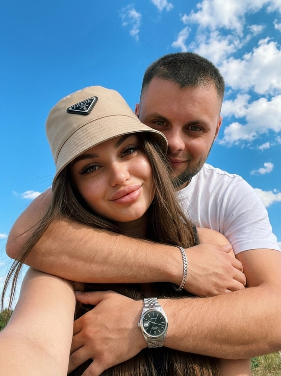 Кинематографичный кадр крупным планом (selfie style), пара в объятиях (классическое "обнимашки сзади") на фоне ярко-голубого неба с облаками.  Женщина на ней бежевая панама с брендовой надписью. Её кожа выглядит сияющей, подсвеченной мягким естественным солнечным светом. Выражение лица спокойное, глубокий взгляд, направленный прямо в камеру. Мужчина стоит строго позади девушки и обнимает её сзади, одна его рука обнимает её за плечи/шею спереди — предплечье лежит поперёк её ключиц, кисть свободно свисает с её плеча, на запястье классические серебряные часы с черным циферблатом. Мужчина в белой футболке, выражение лица доброжелательное, его взгляд прямо в объектив. Ракурс сверху, камера сфокусирована на лице девушки. Высокий контраст, насыщенные цвета, мягкая прорисовка деталей кожи. Эффект пленочной фотографии, легкое зерно, атмосфера летнего дня, эстетика социальных сетей. Телефона не видно в кадре.