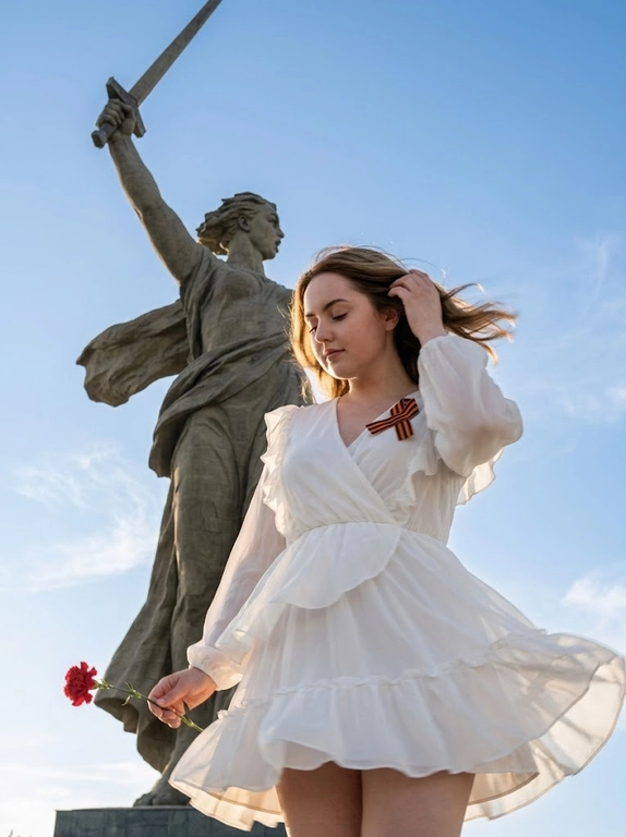 Фотореалистичная съёмка у монумента «Родина-мать зовёт» в
Волгограде. Крупный план, ракурс снизу вверх с небольшим наклоном камеры. Женщина в кадре по пояс, кружится вправо. Лёгкий motion blur на развевающихся волосах, краях ткани и кончиках пальцев. Голова опущена и слегка наклонена, глаза прикрыты, выражение спокойное и сосредоточенное.
Правая рука поднята к голове, заправляет прядь за ухо. Левая рука опущена и держит красную гвоздику. Платье белое летящее с длинными свободными рукавами,
V-образным вырезом, рюшами по декольте и плечам, многослойной воздушной юбкой с оборками, развевается на ветру. На груди приколота Георгиевская лента. На заднем плане монумент «Родина-мать зовёт» - гигантская каменная скульптура женщины с поднятым мечом в правой руке и отведённой в сторону левой, голова повёрнута влево. Ракурс снизу подчёркивает масштаб.
Небо ясное голубое с лёгкими перистыми облаками. Свет естественный солнечный, фронтально-боковой, тёплые блики на коже и ткани, мягкие тени на скульптуре. Композиция: женщина снизу справа, монумент занимает верхнюю часть кадра.
Цветокоррекция нейтральная тёплая, высокая детализация ткани, волос, камня и металла.