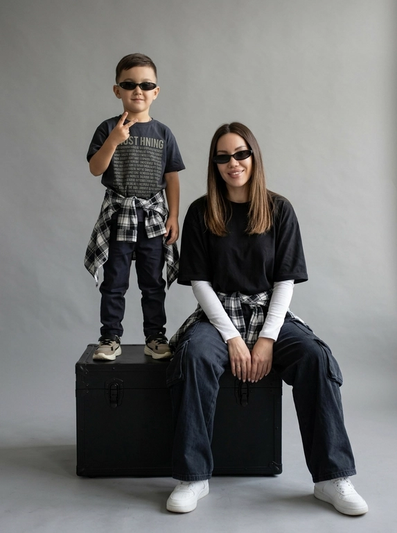 Photorealistic studio portrait photography in minimalistic fashion/editorial aesthetics, static camera at chest level of a seated person, light frontal angle without noticeable perspective deformation; composition with two characters: an model sits on a large matte black cube, body slightly turned towards the camera, knees bent, feet on the floor, the arms are lowered and relaxed in front of the knees, the fingers are neatly extended; the child stands on the same cube behind and to the left, at full height, one hand raised with a "V" gesture, the other lowered, pose confident and playful; both wearing narrow dark sunglasses, gaze directed at the camera; shooting plan — full height for both with a small supply of air above and below, center the center of the frame is shifted to the right to a sitting figure, a standing child balances on the left; the lens is normal/short portrait 50-70 mm, without width; aperture is approximately f/5.6–f/8, moderate depth of field, both characters are sharp, the background is slightly soft; the studio lighting is soft, with one key source on the front left at a level just above the head (softbox), giving smooth transitions and a pronounced but soft shadow against the background on the right; light filling light from the front reduces contrast, shadows are readable; the background is a smooth, light, seamless cyclorama/wall with a noticeable large oval light spot (gradient) in the center, there is a darker vignetting around, a clean minimalistic studio without props; the mood is calm, stylish, slightly retro, with an emphasis on street fashion and family vibe; the expression on the face of the seated model is a soft smile, relaxation; the person standing has a confident neutral facial expression; clothes: the seated model has a dark oversized T-shirt with a large rectangular print on the chest, under it a light longsleeve with long sleeves, wide dark cargo trousers/joggers with pleats at the knees and bottom, a plaid shirt/flannel tied at the waist with a knot, massive light sneakers with a pronounced sole and mesh texture; the child has a dark T-shirt with a large inscription on the chest, dark trousers, a plaid shirt tied at the waist, sneakers in two colors; fabrics with a realistic cotton and flannel texture, noticeable fine folds, natural creases on the knees and in the waist area, matte surfaces without gloss; micro-details: natural skin texture without plastic, neat glare on the lenses of glasses, a clear contour of the shadow from the raised hand on the background, clean floor and soft drop shadow from the cube; high detail, natural sharpness, light grain like a film shot, moderate contrast, soft matte/film look, no HDR, no overexposure and no unnecessary objects.

Какого цвета будут рубашки на поясе?: Чёрно-белый.