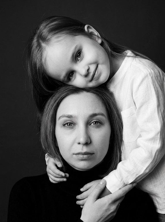 Родитель и ребёнок с внешностью как на приложенных фото, художественная семейная студийная фотосессия. Композиция — вертикальный портрет, крупный план лиц. Родитель внизу смотрит прямо в камеру, лицо фронтально. Ребёнок сверху, голова повернута набок, щека лежит на голове родителя, смотрит в камеру с нежной улыбкой. Рука ребёнка обнимает родителя за шею, видна в кадре, родитель держит руку ребёнка своей рукой.
Одежда: ребёнок в белой льняной рубашке, родитель в чёрной водолазке. 
Фон — тёмно-серый почти чёрный однотонный студийный, создаёт контраст. Освещение — мягкое боковое студийное, создаёт объём и глубину.
Чёрно-белая художественная фотография с выразительными контрастами. Атмосфера нежная, интимная, спокойная.