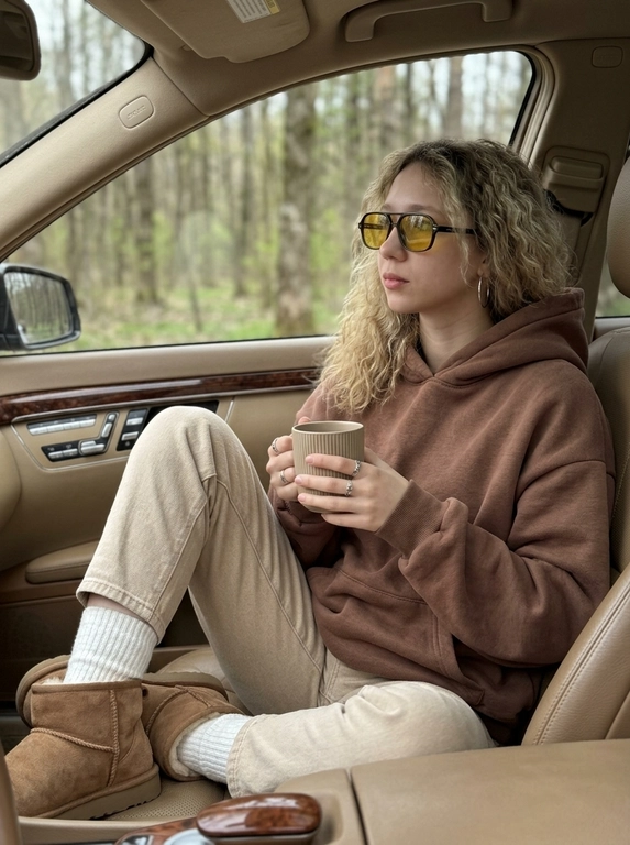 Модель сидит на переднем пассажирском сиденье элегантного автомобиля, повернувшись полубоком к камере, лицо к камере повернуто в три четверти, взгляд направлен вдаль мимо камеры, на ней огромная оверсайз плюшевая толстовка цвета молочного шоколада с капюшоном поверх широких джинсов бежевого цвета, толстые белые носки и уютные уги бежевого цвета с детализированной текстурой. У модели естественный макияж и взъерошенные волосы видны из-под капюшона. На лице надеты солнцезащитные очки в стиле ретро с жёлтыми линзами, в ушах небольшие серьги-кольца. В руках она держит матово-бежевую кофейную чашку с ребристым узором. На пальцах модели крупные серебряные кольца, на которые падает мягкий дневной свет. За окном вид на приглушённый весенний лес с зеленеющими почками на деревьях. Композиция на уровне глаз с небольшой глубиной резкости чётко фокусируется на ней и чашке, в то время как рассеянный дневной свет создаёт резкие тени и блики. Приглушённая палитра серых, чёрных, белых и древесных тонов создаёт уютное весеннее настроение в стиле минимализма благодаря модному редакторскому стилю и гиперреалистичным текстурам.