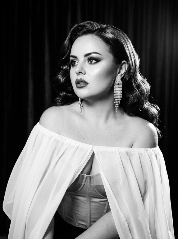 Фотореалистичный контрастный чёрно-белый glamour/editorial портрет в стиле Old Hollywood, вертикальный кадр по грудь. Женщина в 3/4, корпус чуть наклонён вперёд, плечи открыты; голова повернута влево, взгляд вверх/в сторону (не в камеру), губы чуть приоткрыты, выражение драматичное и кинематографичное.  Образ: полупрозрачный белая накидка из шифона с длинными струящимися рукавами, под ним корсет; на ушах крупные серьги со стразами/каскадом. Макияж: классический вечерний glam (в ч/б читается как контраст) — чёткие брови, мягкие smoky тени, тонкая стрелка, длинные ресницы, губы с чётким контуром и сатиновым финишем. Фон: тёмный студийный с мягким рисунком/драпировкой (как бархатные шторы), размытый. Свет: жёсткий key слева спереди (beauty dish) + контровой/rim справа для отделения волос, глубокие тени, яркие блики на скулах и волосах, богатый тональный диапазон (глубокие чёрные и сливочные белые). Камера: 85mm, f/2.0–2.8, ISO 100–200, 1/160, резкость на глазах и губах, фон в мягком боке, лёгкое плёночное зерно, без “пластика”.

Какая будет укладка? : Волосы: объёмные голливудские волны, аккуратная укладка с сильным объёмом и мягкими завитками, блеск волос.