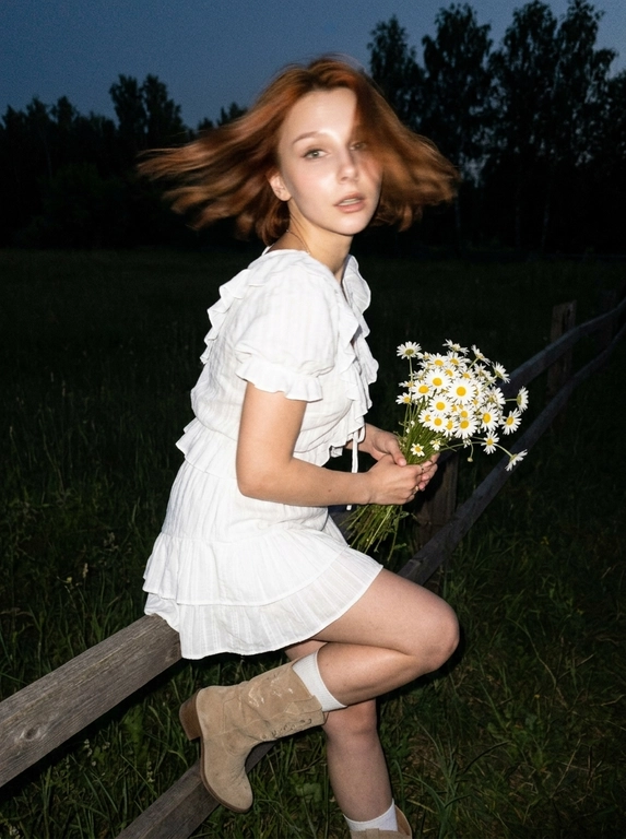 Cinematic candid night shot. iPhone RAW, direct flash, film grain aesthetic.

Girl standing leaning her back and elbows on a wooden fence in a field, distant trees behind her. She suddenly turned her head sharply toward the camera — motion blur from the sharp head turn: hair blur sweeping across the frame, slight blur on face edges, face still recognizable, expression caught mid-movement, alive and unexpected.

hair blown by wind — blur from the sharp turn creates dynamic hair trails across the frame, windswept movement intensifies the motion. Hand holding a bouquet of daisies caught mid-motion.

White short oversized summer blouse with tie detail on chest and ruffles. Matching short skirt with ruffles, same fabric. Beige short suede cowboy boots, white socks slightly visible.

Flawless makeup, dusty rose lips, bronzed tanned skin. Direct camera flash hits face and upper body — skin glowing and shiny, wet oil-skin effect, highlights on cheekbones, nose, forehead. Deep shadows behind her, background falls into dark night.

Dark night, no ambient light except flash. Wooden fence and distant tree silhouettes barely visible in the darkness. Flash creates strong contrast — figure brightly lit, everything behind fades to black.

Cinematic film grain, high ISO sensor noise, muted dark palette, no beauty filter, no plastic skin, no AI look, no watermark, no text.

--ar 9:16 --raw --style motion 50