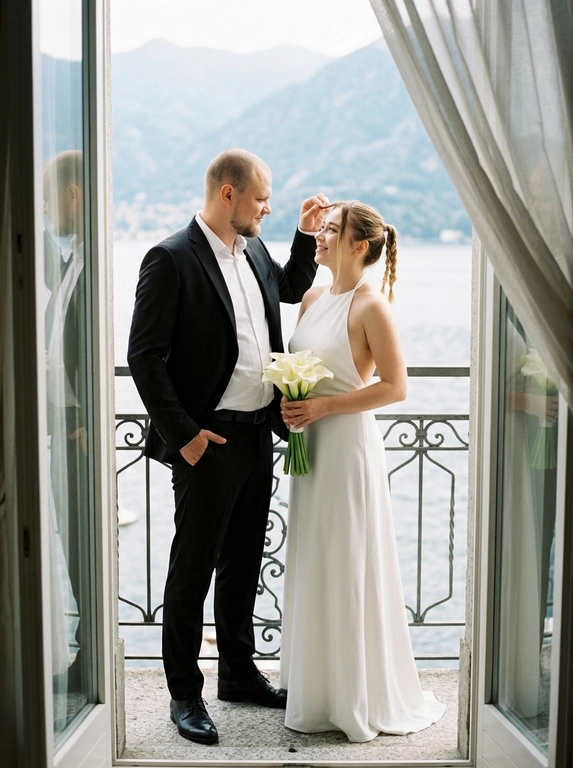 Фотореалистичный bridal editorial, полный рост. Пара стоит на узком балконе у открытой стеклянной двери. Мужчина слева, корпус в 3/4 вправо к женщине, одна рука в кармане брюк, другой рукой мягко касается её волос у лба, голова чуть наклонена к ней, взгляд на женщину, лёгкая нежная улыбка. Женщина справа, стоит очень близко, корпус в 3/4 влево, голова поднята к мужчине, взгляд снизу вверх на его лицо. В обеих руках прижат к животу маленький аккуратный букет из 11 этих цветов: бутоны цветов собраны плотно, стебли строго вниз, букет вертикальный, не у бедра, не у груди. Мужчина в чёрном костюме и белой рубашке без галстука. Женщина в длинном белом платье-халтер с открытой спиной. Фон: кованые перила, озеро, горы, светлые шторы. Мягкий естественный дневной свет, 50mm, лёгкое зерно.

Из каких цветов будет букет невесты?: Белая лилия калла