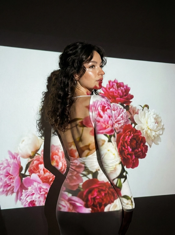 Кинематографический эффект с добавлением зерна. Ультрареалистичный портрет женщины, стоящей на фоне белой стены: её тело немного повёрнуто в сторону.  Фото с нижнего ракурса, фото со спины девушки. Макияж лёгкий: хайлайтер, длинные ресницы. На ней белое однотонное платье по фигуре, с открытой спиной и длинными рукавами. Она не смотрит в камеру. Освещение мягкое, с лёгким акцентом на её лицо, создающее нежные тени и подчёркивающее черты. Вокруг темно — светит только проектор с изображением ярких пионов прямо на девушку и на стену за ней. Только один источник света — проектор.


Какая будет прическа?: Длинные волосы, собранные в мягкие локоны