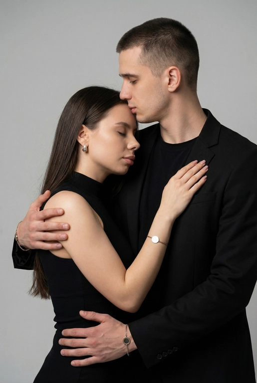   Мужчина и женщина позируют в студии в объятиях. Мужчина стоит позади женщины, обнимая её обеими руками; его тело повернуто к ней, голова слегка наклонена вниз, губы касаются её лба. Женщина немного отклоняет голову назад к его плечу, её глаза закрыты. Кадр — средний, от талии до головы, снят на уровне глаз. Композиция плотная, акцентирует линии рук и соприкосновение лиц. Женщина стоит, прямо обращённая к камере.
На ней облегающий чёрный топ без рукавов с высоким воротом, из плотной матовой ткани.
Мужчина одет в чёрную футболку и чёрную неброскую куртку. На обоих запястьях — тонкие металлические браслеты с минималистичными круглыми подвесками, а женщина носит небольшие светлые металлические серьги с гладкой поверхностью.
Обстановка минималистичная: однотонный светло-серый фон без текстуры.
Освещение — мягкий студийный свет: рассеянный фронтальный и боковой свет создаёт плавные переходы света и тени на лицах и руках. Тени мягкие, средней глубины. Контраст умеренный, без жёстких бликов. Свет равномерно распределён по фигурам, фон подсвечен мягко, без выраженных градиентов.
Съёмка на портретный объектив 85 мм, малая глубина резкости с чётким фокусом на лицах и руках; фон слегка размыт, текстуры кожи и ткани высоко детализированы. Это минималистичный студийный портрет, высокодетализированный и фотореалистичный.
Минималистичное романтическое студийное настроение, нейтральные тёплые тона кожи, низкая–средняя насыщенность, мягкое рассеянное освещение, средний контраст с нежными тенями, плавные переходы бликов, естественная передача кожи, чистый светлый фон, тонкий тональный баланс, современная мягкая редакционная цветокоррекция. Не изменять мою прическу