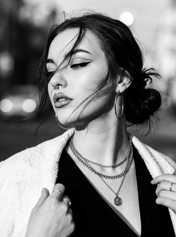 Элегантность в движении (B&W)
Максимально реалистичная фотография, эстетика высокого глянца и кинематографичного нуара.

Тип кадра: Крупный план (close-up portrait). женщина в профиль, голова слегка наклонена вниз. губы слегка приоткрыты, поза выражает сдержанную уверенность и стиль.

Макияж: Графичные черные стрелки, подчеркивающие разрез глаз, четкий контур губ, матовый тон. В монохромном исполнении акцент на контрасте света и тени.

Одежда: Черный топ с глубоким V-образным вырезом, на плечи накинут белый структурированный жакет. массивные серьги-кольца, многослойные тонкие цепочки с кулоном на шее.

Локация: Минималистичный городской фон, залитый светом, создающий мягкое боке.

Освещение: Контрастное черно-белое освещение (high contrast B&W). Яркий боковой свет подчеркивает линию челюсти, скулы и изгиб шеи, создавая глубокие тени.

Камера: 85мм, объектив с высокой светосилой, имитация мелкозернистой черно-белой пленки.

Глубина резкости: Лицо и украшения в резком фокусе; волосы на переднем плане и жакет на плече уходят в мягкое размытие.

Кожа: Реалистичная текстура, видны поры, микрорельеф на шее и ключицах, отсутствие ретуши.

Детали: Блики на цепочки, четкие тени от прядей волос на лице, текстура ткани жакета.

Атмосфера: Загадочная, дорогая, изысканная.

Без: ИИ-эффектов, пластиковой кожи, пересветов, HDR, неестественных цветов, хроматических аберраций.

Какая будет прическа?:  Собраны в небрежный низкий пучок; несколько тонких, хаотичных прядей выбились и закрывают часть лица, создавая эффект динамики и ветра.

Очки: На ней нет очков