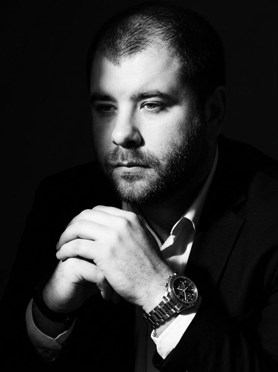 A hyper-realistic 8K cinematic portrait of a uploaded person,
captured in a dramatic Chiaroscuro style. The subject is seated at a
three-quarter angle, leaning slightly forward with a relaxed yet
commanding posture. His face is turned away from the lens, with
one side of his face clearly illuminated while the other is
completely lost in deep, velvety black shadows. His expression is
contemplative, not looking at the camera. The pose is precise: his
hands are clasped together near his chest; one hand is held
straight with fingers slightly tilted, while the other hand is
positioned so only the fingers are visible, resting over the first. He
wears a luxury black chronograph watch with a detailed metal link
bracelet, and a beaded bracelet. His attire is a sharp
black suit jacket over a white dress shirt with the top buttons
open, showing refined fabric textures. The background is a solid,
seamless black. The lighting is sharp and directional, highlighting
the intricate details of his hair,
the moisture in his eyes, and the glint off the watch face.
Cinematic black-and-white color grade with rich contrast and
smooth, natural skin transitions. Shot on 85mm lens, f/1.8, high-
resolution commercial photography