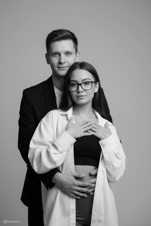 Черно-белый портрет мужчины и женщины в стиле фэшн фотосессии в фотостудии. Не изменяйте лица. Фон нейтральный, однотонный, светло-серый. Атмосфера интимная и спокойная. На переднем плане женщина. Вся композиция выполнена в приглушенной монохромной гамме со студийным освещением, которое подчеркивает текстуру кожи и одежды. Её руки лежат на груди мужчины. Мужчина обнимает женщину сзади, положив свою руку поверх её руки. Она одета в черный топ, белую оверсайз рубашку. Мужчина одет в  классическом черном пиджаке и белой футболке.