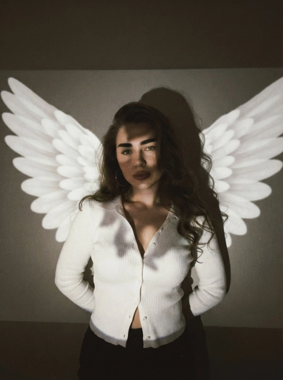 A raw vintage low-light photograph taken in a dark room with a projector. Medium shot, framed from the hips to slightly above the head. Camera directly in front of the woman at approximately chest level, almost straight-on. She stands centered very close to a plain wall.

POSE: her torso faces directly toward the camera. BOTH ARMS are completely placed behind her back, with neither hand visible. Her shoulders are gently pulled backward because of the position of her arms, while her upper body leans very slightly toward the camera. Head subtly tilted to one side and lowered, direct calm gaze into the lens.

She wears a fitted white ribbed LONG-SLEEVE button-front top. The fabric closely follows the body. Several buttons at the upper chest are casually left undone, creating a narrow deep V opening; the lowest buttons near the waist are also slightly undone, while the middle section remains buttoned. Long fitted sleeves extend fully to the wrists. Large thin hoop earrings.

A projector casts a flat image of enormous white angel wings onto the wall behind her. The wings spread horizontally from behind both shoulders almost to the edges of the frame. They are projected light, NOT physical wings. Large rounded individual feathers are clearly visible.

The projector simultaneously creates uneven patches of bright white light across the long-sleeve top, while the face, neck and upper chest remain partially covered by deep irregular shadows. A large dark silhouette of her head appears on the wall.

Very dim exposure, warm gray-brown shadows, creamy projector whites, slightly blown highlights, crushed blacks, soft focus, coarse grain, visible digital noise and slight pixelation. Authentic imperfect late-1990s / early-2000s amateur photograph, low-resolution nostalgic camera quality, no polished studio look.

Причёска: Не менять прическу

Доп. характеристики: Не менять черты лица