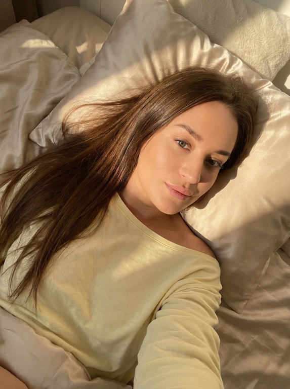   Создай портрет, не меняя черты лица. Девушка лежит на кровати делая селфи от первого лица, она в нежно желтом лонгсливе, спущенном с плеча
Волосы цвет как на фото распущенные блестящие лежат на подушке, голова полубоком.
Постель  светло бежевая сатиновая, фото в золотой час
Солнце красиво освещает её лицо, её безупречный макияж.
