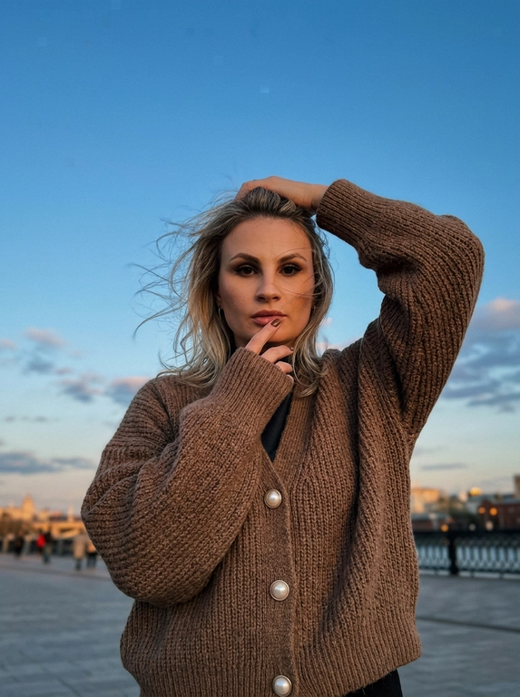 Сверхреалистичная уличная fashion-фотография на iPhone 17 Pro Max, основная камера 1x, 24 мм. Средний портрет примерно до середины бёдер, камера на уровне груди, женщина по центру кадра, над головой много открытого вечернего неба.

Женщина стоит на открытом пространстве, корпус почти фронтально к камере. Сильный ветер активно развевает волосы в стороны, отдельные тонкие пряди хаотично проходят перед лицом. Одна рука поднята над головой и лежит на макушке, локоть направлен в сторону; вторая рука согнута перед грудью, длинный рукав частично закрывает кисть, пальцы находятся возле губ. Взгляд прямо в объектив, губы сомкнуты, выражение уверенное, без улыбки.

На ней объёмный коричневый вязаный кардиган oversize с жемчужными пуговицами, длинными свободными рукавами и выраженной мягкой фактурой крупной вязки. Кардиган свободно собирается складками на теле.

Макияж выразительный, но естественный: кожа с лёгким загаром и натуральной текстурой, оформленные брови, длинные густые ресницы, мягкая чёрная подводка, лёгкий бронзер и румянец, пудрово-розовые губы, ногти коричневые.

Фон минималистичный: красивая вечерняя московская улица. Большую часть кадра занимает чистое голубое вечернее небо с небольшими мягкими светлыми облаками.

Тёплые золотистые блики на лице и волосах, мягкие естественные тени. Цветокоррекция слегка плёночная: приглушённые натуральные оттенки, тёплая кожа, немного пониженная экспозиция, умеренный контраст, лёгкая зернистость. Волосы должны двигаться физически естественно от сильного ветра. Без студийного света, пластиковой кожи, сильного боке, CGI и AI-эффекта. Кадр будто сделан в спешке и размыт на 2%.