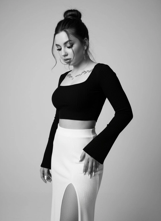 Профессиональная фэшн-фотография в чёрно-белом стиле, молодая привлекательная женщина стоит в полупрофиль на однотонном светло-сером фоне, чёрный облегающий кроп-топ с длинными рукавами и квадратным вырезом, ткань эластичная резинка, обнажает живот, белая юбка-макси из той же рифлёной ткани с высокой посадкой и глубоким V-образным разрезом спереди, открывающим часть живота и бёдер, рукава топа расширены к низу — форма bell sleeves, волосы собраны в высокий пучок на макушке, несколько прядей свободно падают на лицо и шею, большие круглые серьги-кольца из металла. Внешность:Густые, длинные накладные ресницы (false lashes), создающие эффект «кошачьего глаза». Брови идеально уложены, ламинированы, имеют четкую форму и темный цвет. Легкий румянец.Светоотражающий хайлайт на щеках Губы с блеском.Ногти длинные молочного цвета квадратные. Фигура песочные часы большая грудь четвёртого размера и широкие бёдра. Промт: Leronitg губы слегка приоткрыты, модель стоит в профиль, корпус слегка повёрнут к камере, голова опущена вниз, взгляд направлен вниз, левая рука согнута в локте, ладонь лежит на бедре или внутри разреза юбки, правая рука опущена вниз, пальцы слегка согнуты, задумчивое сосредоточенное выражение лица, мягкое студийное освещение спереди и слева, нет жёстких теней, блик на лбу, носу, щеке, ключицах, свет падает так, чтобы подчеркнуть объём груди, талии и бёдер, чёрно-белый фильтр с высоким контрастом, тени глубокие, света яркие, профессиональная ретушь кожи, эффект скульптурной красоты, стиль арт-фотографии или высокой моды, размытие фона отсутствует, вертикальный формат 3:4, средний план от головы до колен, композиция центрирована на фигуре, атмосфера загадочности и чувственности, современный минимализм, монохромная палитра, фотореализм, 8K качество, эмоциональный портрет, внутренняя сила и уверенность