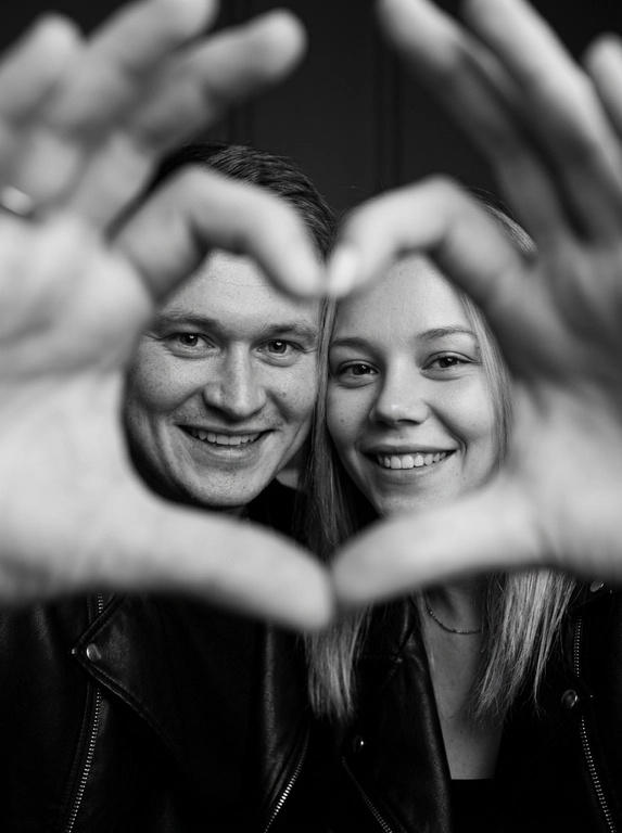 Ultra-realistic editorial photography of a smiling couple in a romantic and cinematic composition. The couple is centered in the background of the image, looking directly at the camera with a spontaneous, happy, and natural expression. In the foreground, extremely close to the lens and slightly out of focus, the couple’s hands form a heart shape perfectly framing their faces.

The composition creates strong visual depth, with sharp focus on the eyes and faces inside the heart formed by the hands. The hands occupy a large portion of the foreground, artistically blurred around the edges, creating a sophisticated cinematic effect.

Intimate, emotional, and elegant aesthetic inspired by modern perfume campaigns and premium lifestyle editorials. Soft studio lighting with delicate contrast and natural shadows. Minimalistic dark blurred background.

The couple wears minimalistic black leather jackets and sophisticated dark clothing. Genuine expressions, strong emotional connection, contemporary romantic atmosphere. Hyper-realistic skin texture, visible pores, detailed hair strands, authentic human appearance.

Black and white photography, rich tones, deep blacks, and soft highlights. 85mm lens, shallow depth of field, cinematic portrait photography, luxury editorial style, ultra detailed, hyper realistic, 8K.