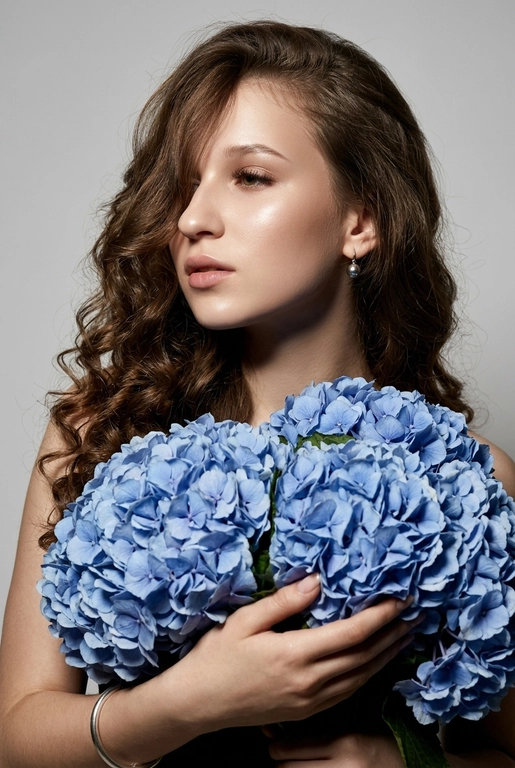 Создайте ультрареалистичный портрет, не меняя черты лица, крупным планом 3/4, с мягкой, освещённой кожей, атласной отделкой и естественным блеском, где женщина изображена в профиле. Спокойное и чувственное выражение лица, губы слегка полуоткрыты. Она носит серебряные серьги-шары и простой серебряный браслет на запястье.
Фон нейтральный, в светло-сером тоне, с тёмным, целенаправленным низким угловым освещением, которое подчёркивает контур лица, текстуру кожи и объём волос. Стиль редактирования — высокая мода, чёткий фокус 8K, реалистичная текстура, изысканная и элегантная атмосфера.


Длинные волнистые волосы покрывают часть лица объёмными локонами, выпадающими на лицо и плечо. 

Цветы: Держит в руках огромный букет голубых гортензий