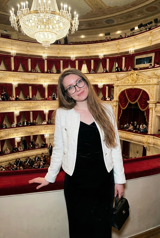 Я стою в ложе Большого театра, на рядов лож и большой хрустальной люстры. В ложах сидят люди, висят красные бархатные занавески. Камера на уровне глаз.  Съёмка выглядит как фото, сделанное на iPhone: естественный свет, лёгкое зерно, минимальная обработка, живой момент без позирования. Взгляд в камеру, легкая улыбка. Голова немного наклонена вбок. Волосы сияют, макияж естественный, губы глянцевые. Одна рука лежит на бортике ложа, во второй - маленькая черная сумочка. Приглушенный свет. На мне чёрное бархатное вечернее платье без рукавов по фигуре и белый жакет в стиле Channel. 