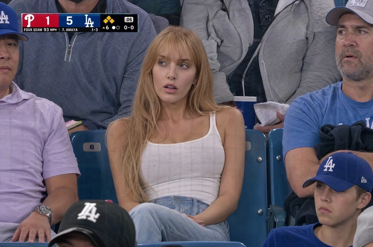 Person sitting in the crowd at a professional baseball game (or it could be any other match that I will write below), filmed from far away by a live broadcast camera. 
Seated in blue stadium chairs, slightly leaned back, looking off to the side with a surprised caught-on-camera expression, lips slightly parted.

Какая будет одежда? : Поменяй одежду на топ с открытыми руками и джинсы, мои волосы по пояс