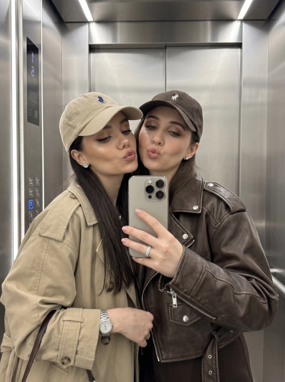 Не меняя черты лица людей с добавленного фото. Ультра-реалистичное фото в зеркало на смартфон iPhone 16 Pro Max, цвета титан, двух женщин, позирующих близко друг к другу в современном лифте. Две женщины, стоящие плечом к плечу, лица очень близко к камере, обе делают игривые выражения губами в виде поцелуя. Женщина слева: бежевого цвета бейсболка Ralph Lauren с маленьким вышитым логотипом поло, oversized бежевый тренч-жакет с большими пуговицами и широкими рукавами, серёжки-гвоздики с бриллиантами, на запястье серебряные часы с большим циферблатом, на пальце кольцо из страз в виде дорожки, макияж: хайлайтер, ресницы лисий эффект. Женщина справа: тёмно-коричневая бейсболка Ralph Lauren с вышитым логотипом, oversized шоколадно-коричневая укороченная кожаная куртка с серебряной фурнитурой и погонами на плечах, серёжки-гвоздики с бриллиантами, мягкий глэм-макияж с выраженными скулами, подводкой для глаз и глянцевыми губами. Поза и композиция: обе женщины слегка наклоняют головы друг к другу.Окружение: современный просторный лифт. Освещение: мягкое внутреннее освещение, равномерно освещающее лица.Фото в зеркало на iPhone 16 Pro Max в стиле высокой реалистичности.