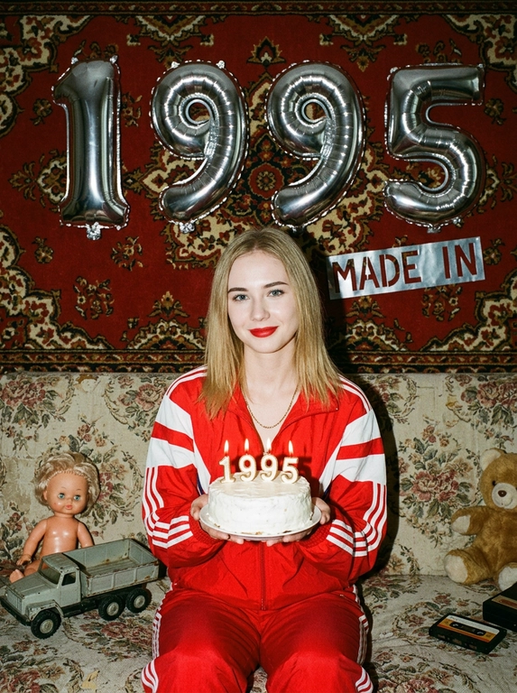 Создай крупный портрет, не меняя черты лица. На фотографии модель в стиле «лихие 90-е» в России. Модель сидит на старом диване с цветочной обивкой на фоне красного советского ковра на стене и смотрит в камеру с лёгкой улыбкой. На ковре висят большие надувные серебряные фольгированные шары в форме цифр и надпись «MADE IN». На модели классический красный спортивный костюм того времени и кроссовки. В руках маленький праздничный белый торт со свечами. Рядом на диване лежат игрушки: советские игрушки и старая советская кукла. Фото со вспышкой. Освещение мягкое, создающее атмосферу ностальгии. Высокое разрешение, реализм. Модель в олдскульном спортивном костюме сидит расслабленно, с лёгкой улыбкой. Ретро-вайб, приглушённые цвета, зернистость плёнки, детализированные текстуры ткани и шерстяного ковра.

Какая будет цифра?: Цифра 1995.