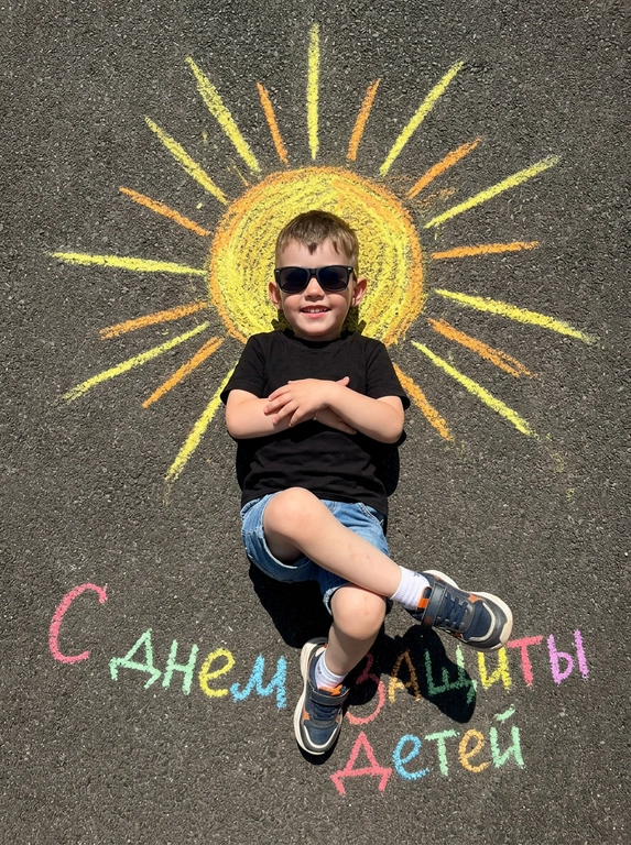Top-down shot of a child lying on dark grainy asphalt. A bright chalk-drawn sun with radiant rays is drawn above the child’s head. The child is wearing black sunglasses, a black t-shirt, denim shorts, and sneakers with white socks. Arms crossed on chest, legs bent and crossed at ankles. Vibrant chalk sun texture in yellow, orange, and warm golden tones. Harsh midday sunlight, sharp shadows, high contrast. Summery playful atmosphere. On the asphalt below, colorful chalk lettering reads "С днем защиты детей". Photorealistic, 8k, highly detailed texture.

Доп. характеристики: Не меня очертание лица