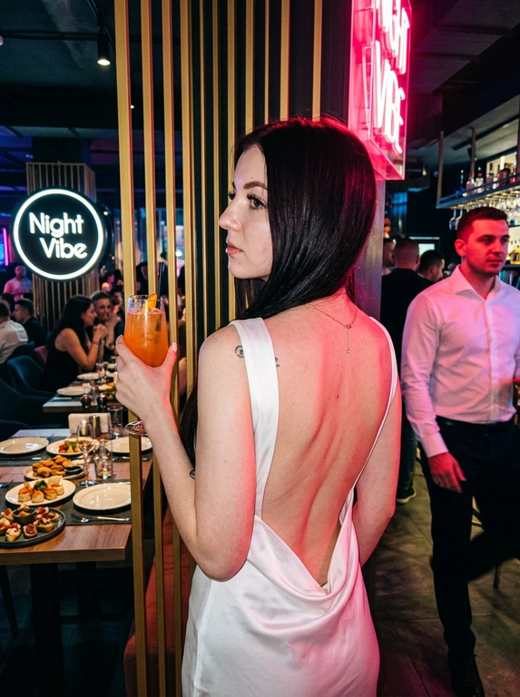 Ресторан. На этой фотографии запечатлена женщина в обстановке ночного ресторана, снимок сделан со спины в пол-оборота, лицо в профиль, что создает динамичный и выразительный образ. На женщине белое атласное платье с открытой спиной, этот наряд подчеркивает фигуру и соответствует вечернему дресс-коду. В правой руке она держит высокий стакан с напитком оранжевого цвета (возможно, коктейль), на шее можно заметить золотое украшение. Макияж — длинные ресницы с эффектом «лучики», губы накрашены блеском. Маникюр короткий квадрат жемчужного цвета. Женщина стоит у барной стойки или декоративной перегородки с вертикальными золотистыми рейками, слева на фоне видны накрытые столы с закусками и тарелками, а также круглая вывеска с надписью Night Vibe (вероятно, название заведения). В помещении приглушенный клубный свет, на фигуру ложится яркий розово-красный блик от неонового освещения или прожектора, что добавляет кадру сочности и атмосферности ночной жизни. Справа в кадр частично попал другой человек в белой рубашке, что подчеркивает многолюдность и социальный характер обстановки. Это классический пример фотографии для социальных сетей, передающий эстетику ночного города и отдыха, эстетика Instagram, черты лица полностью сохранены как на исходном фото.