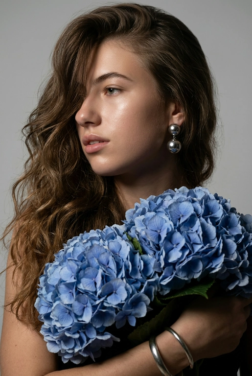 Создайте ультрареалистичный портрет, не меняя черты лица, крупным планом 3/4, с мягкой, освещённой кожей, атласной отделкой и естественным блеском, где женщина изображена в профиле. Спокойное и чувственное выражение лица, губы слегка полуоткрыты. Она носит серебряные серьги-шары и простой серебряный браслет на запястье.
Фон нейтральный, в светло-сером тоне, с тёмным, целенаправленным низким угловым освещением, которое подчёркивает контур лица, текстуру кожи и объём волос. Стиль редактирования — высокая мода, чёткий фокус 8K, реалистичная текстура, изысканная и элегантная атмосфера.


Длинные волнистые волосы покрывают часть лица объёмными локонами, выпадающими на лицо и плечо. 

Цветы: Держит в руках огромный букет голубых гортензий