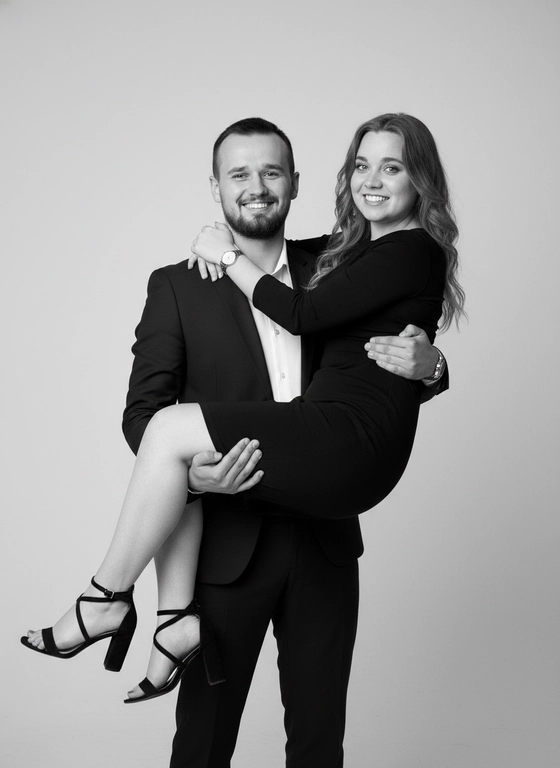 
Чёрно-белое студийное фото, изображающее пару. Мужчина одет в классический тёмный костюм с пиджаком на одной пуговице и белой рубашкой с расстёгнутым воротником. На ногах тёмные брюки. На запястье - часы. На женщине надето облегающее короткое тёмное платье и чёрные босоножки на каблуке с тонкими ремешками.

Мужчина держит женщину на руках. Она наклонена, смотрит в камеру и улыбается. Он также смотрит в камеру с улыбкой.

Фон светлый, однотонный. Стиль фото - элегантный и непринужденный. Атмосфера радостная и романтическая.