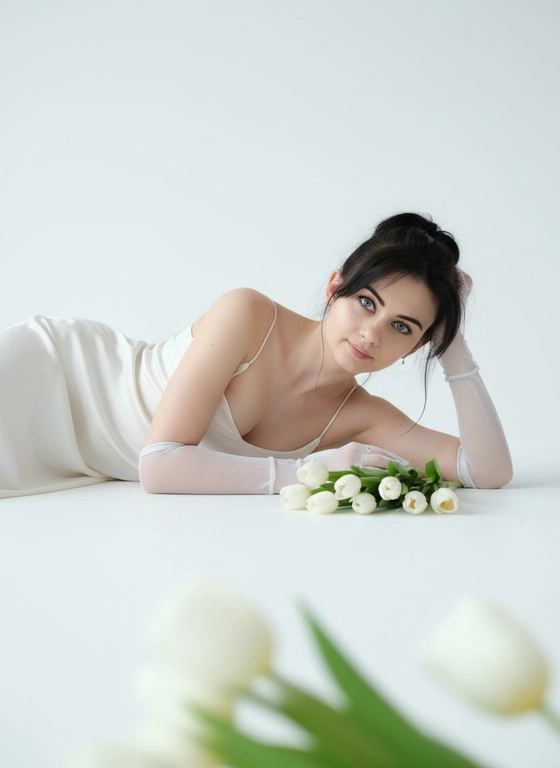 Эта женщина лежит на белой поверхности на боку, голова на согнутой руке. Не меняй ее черты лица, возраст, волосы. Крупный план. На ней молочное атласное платье на тонких бретельках и полупрозрачные белые сетчатые перчатки. Перед лицом лежит букет белых тюльпанов, цветы на переднем плане слегка размыты. Камера на уровне глаз, крупный портрет. Взгляд в камеру, спокойное выражение лица. Мягкий рассеянный дневной свет, белая студия, чистый белый фон. Реалистично, высокая детализация кожи, shallow depth of field.