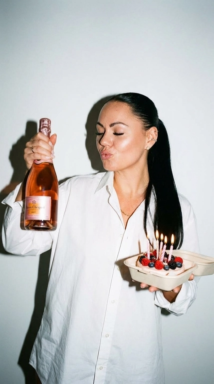 Ультрареалистичная фотография в эстетике Party Girl / Birthday Aesthetic, стилизация под случайный снимок на компактную цифровую камеру начала 2000-х.
 девушка празднует день рождения. На ней свободная белая рубашка оверсайз на пуговицах. В одной руке она высоко держит бутылку розового шампанского, в другой — небольшой бенто-торт со светлыми свечами и свежими ягодами.

Глаза закрыты, на лице игривое выражение с лёгким «поцелуйным» жестом губ. Не меняй прическу модели.

Съёмка со вспышкой прямо в камеру на фоне простой молочно-белой стены. Жёсткий свет создаёт характерную чёткую тень за фигурой, яркие блики на коже, волосах, бутылке и глазури торта. Атмосфера спонтанного домашнего праздничного снимка: немного несовершенная композиция, лёгкая зернистость, естественная текстура кожи, реалистичные отражения и мелкие детали.

Стиль: 2000s digital camera, lo-fi party aesthetic, candid snapshot, hard flash, authentic lifestyle photography, natural skin texture, realistic colors, high detail, 4K.

Без: студийной постановки, мягкого идеального света, пластиковой кожи, чрезмерной ретуши, CGI, мультяшности, искажённых рук и пальцев, лишних пальцев, деформированной бутылки или торта, артефактов текста, логотипов и водяных знаков.