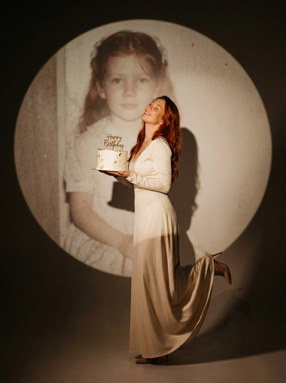 Create ultra-realistic cinematic birthday photoshoot, elegant studio with a dark background and a large circular spotlight projection on the wall. Inside the spotlight, a faded vintage childhood photo of the same girl(the reference image 2nd given)appears as a nostalgic memory, slightly transparent with old photo texture and soft blur. In the foreground, the present-day version of the same model (the reference image 1st given)  holding a white birthday cake decorated with gold accents with a “Happy Birthday". 

If the model is a woman, then she is wearing a cream long gown. If the model is a man, he is wearing a suit.

her head tilted back, eyes closed, natural smile,  cinematic shadow cast on the wall, warm soft spotlight lighting.

If the model was a woman, she additionally lifted her leg in an elegant pose.