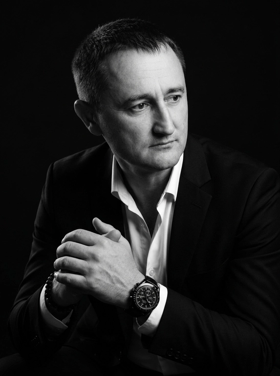 A hyper-realistic 8K cinematic portrait of a uploaded person,
captured in a dramatic Chiaroscuro style. The subject is seated at a
three-quarter angle, leaning slightly forward with a relaxed yet
commanding posture. His face is turned away from the lens, with
one side of his face clearly illuminated while the other is
completely lost in deep, velvety black shadows. His expression is
contemplative, not looking at the camera. The pose is precise: his
hands are clasped together near his chest; one hand is held
straight with fingers slightly tilted, while the other hand is
positioned so only the fingers are visible, resting over the first. He
wears a luxury black chronograph watch with a detailed metal link
bracelet, and a beaded bracelet. His attire is a sharp
black suit jacket over a white dress shirt with the top buttons
open, showing refined fabric textures. The background is a solid,
seamless black. The lighting is sharp and directional, highlighting
the intricate details of his hair,
the moisture in his eyes, and the glint off the watch face.
Cinematic black-and-white color grade with rich contrast and
smooth, natural skin transitions. Shot on 85mm lens, f/1.8, high-
resolution commercial photography